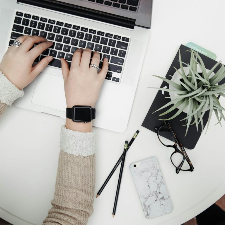 Hände tippen auf einer Tastatur, umgeben von Notizblock, Telefon und Pflanze.