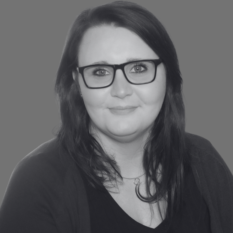 Porträt einer Person mit Brille und langen, dunklen Haaren vor einem grauen Hintergrund.